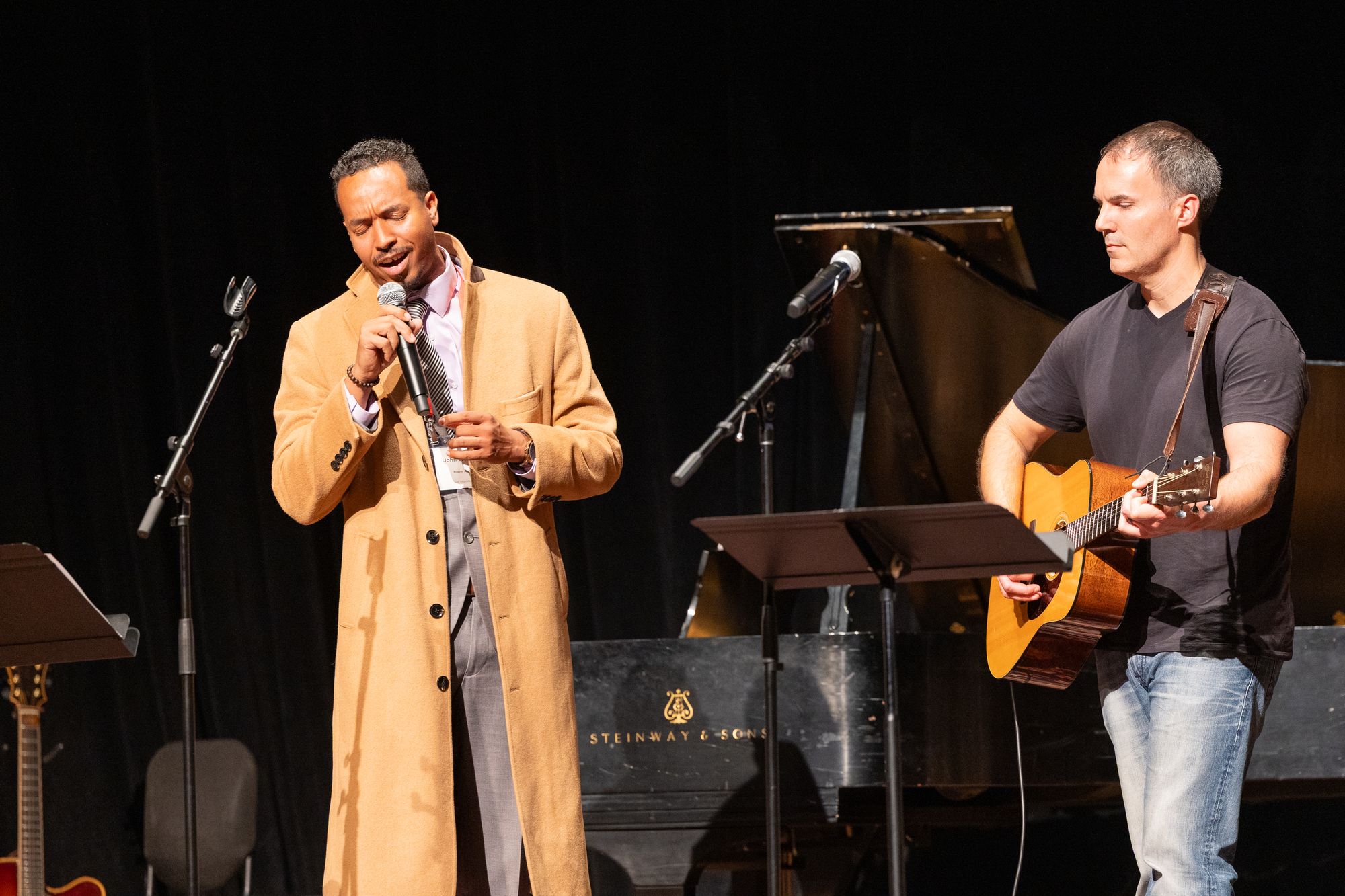John Wood Jr. singing on stage, accompanied by a guitarist, with a grand piano behind them
